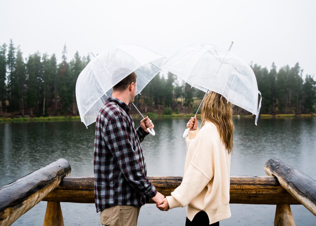 married couple standing in the rain with umbrellas at sprague lake during their rocky mountain national park engagement photos