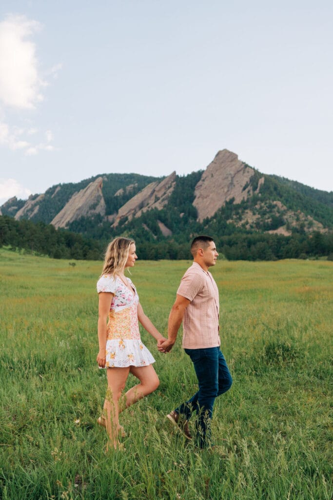 Couple holding hands in grassy field, mountains behind.