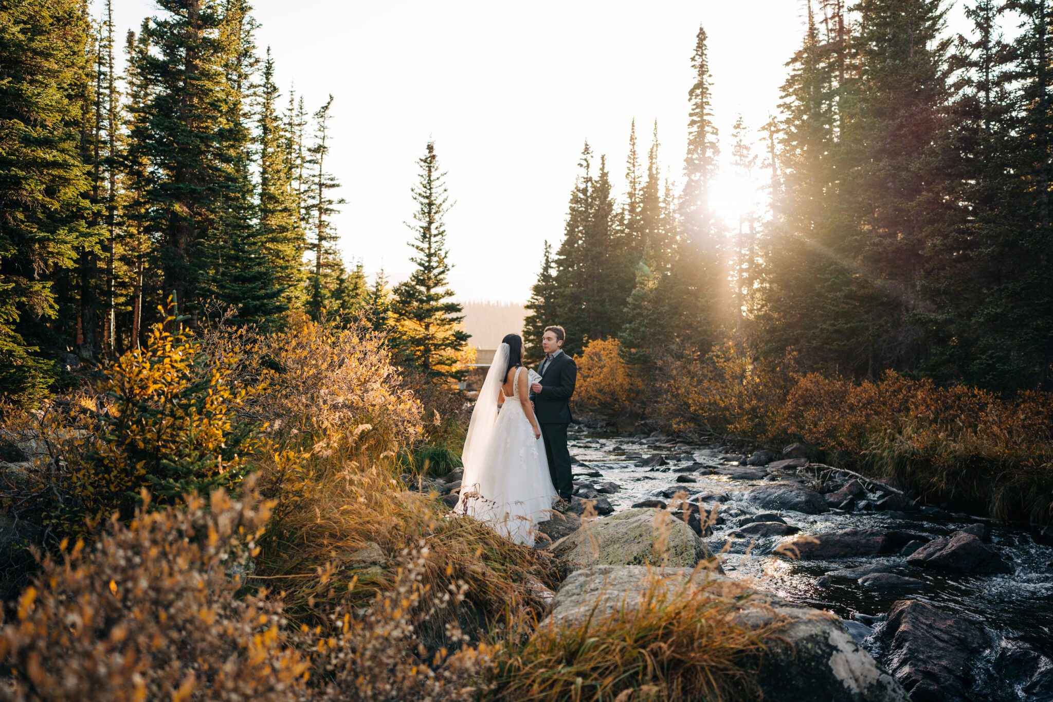 A bride and groom stand together beside a rocky creek at Brainard Lake, surrounded by tall evergreen trees and autumn foliage, with sunlight streaming through the forest—perfect inspiration for an elopement.