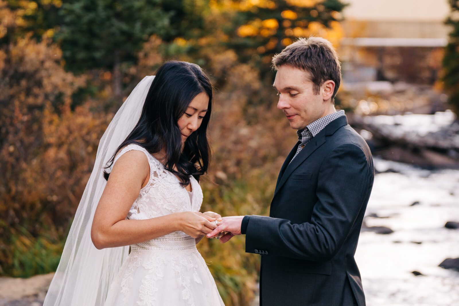 A bride in a white dress places a ring on the grooms finger during an outdoor wedding ceremony by a stream, surrounded by autumn trees and greenery.