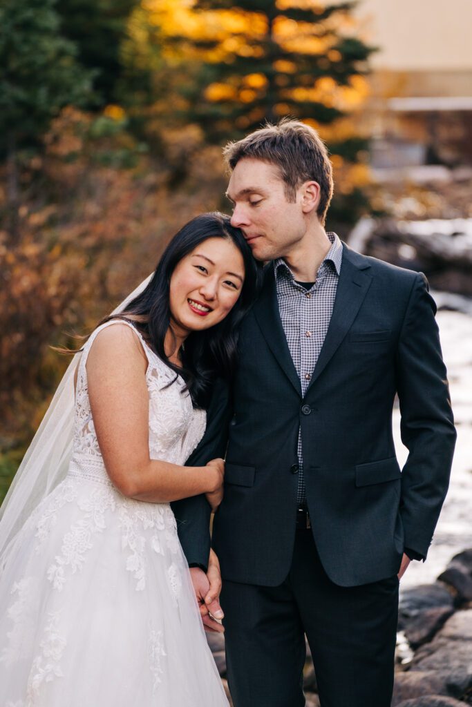 A bride in a white lace dress leans on her smiling groom in a dark suit. They hold hands outdoors by a stream, with trees and autumn foliage in the background. The groom gently kisses her head.