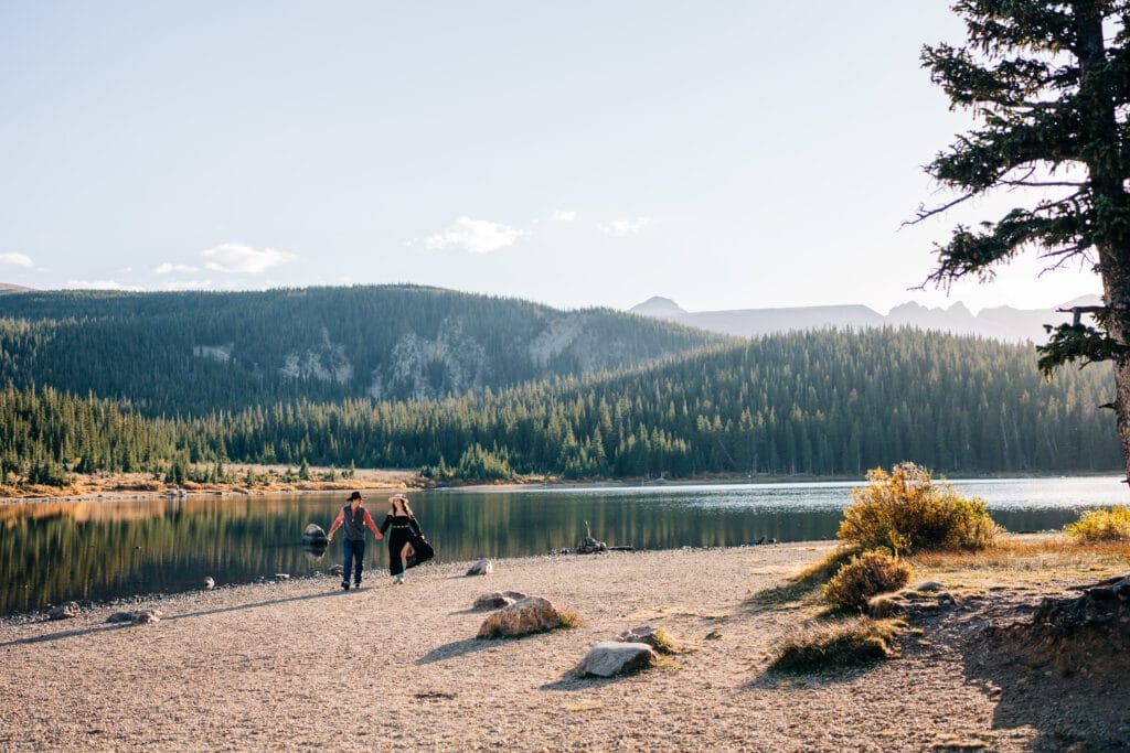 Three people walk along a lakeshore surrounded by trees and mountains under a clear sky, with sunlight reflecting off the lake and a pine tree visible on the right.