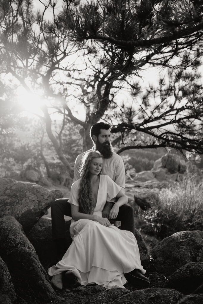 A couple sits together on rocks under a tree, bathed in sunlight. The woman in a flowing dress sits in front, leaning back against the man, who has a beard. Both appear calm and serene in the natural setting. Black and white photo.