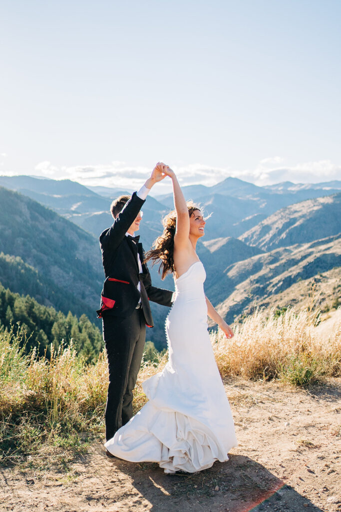 A bride and groom dance joyfully outdoors on a sunny day, surrounded by mountains and grassy hills, with the bride in a white dress and the groom in a black suit.