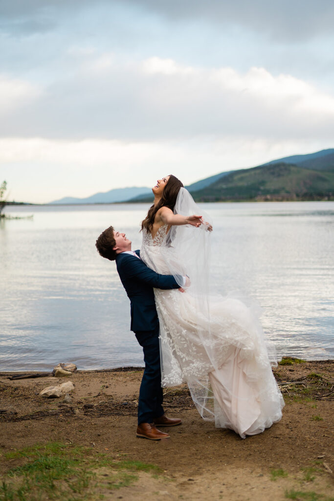 A groom in a navy suit lifts his laughing bride in a lace wedding dress by a lake, with mountains and cloudy sky in the background. The couple appears joyful and carefree outdoors.