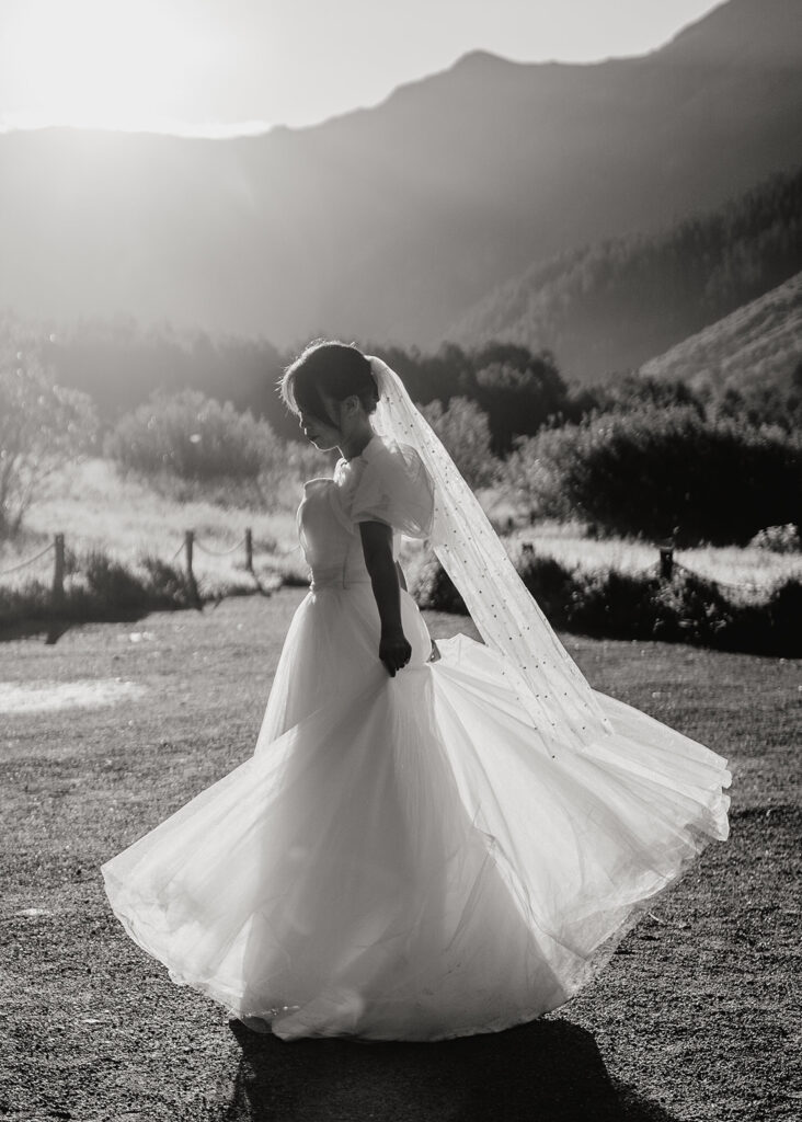 A bride in a flowing wedding dress and veil twirls outdoors, backlit by the sun with mountains and trees in the background. The image is in black and white.