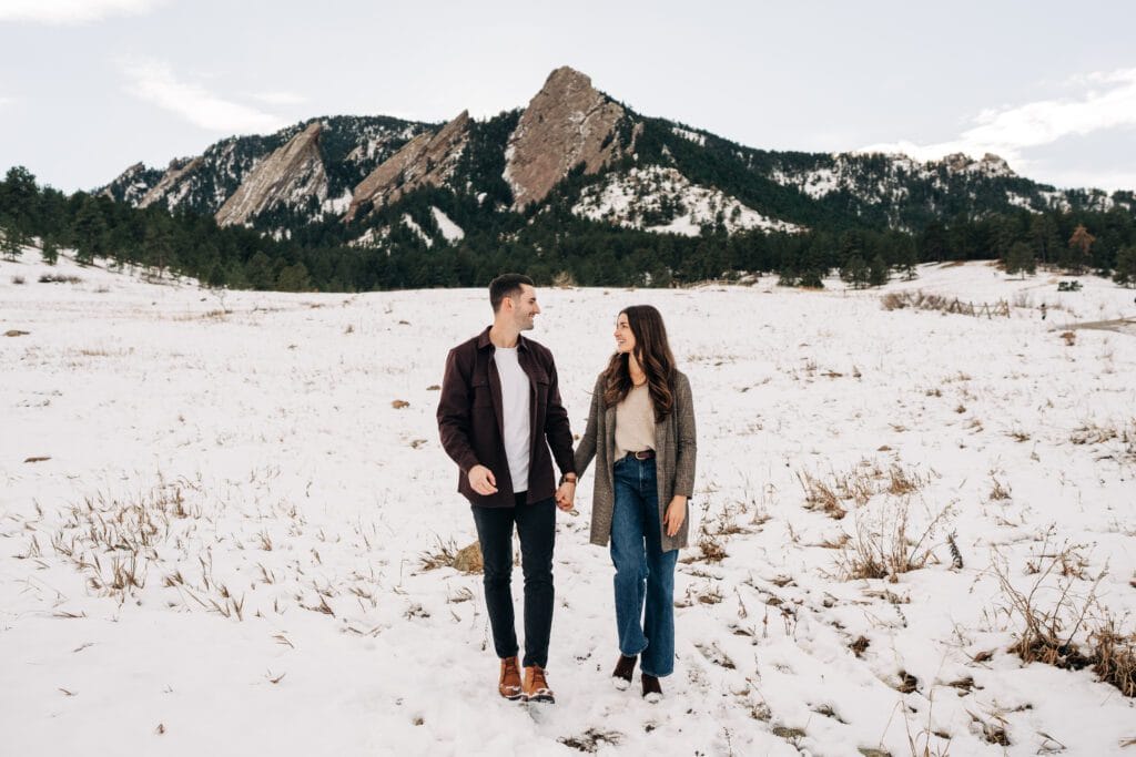 Engaged couple holding hands walking in a snowy field with the Boulder, Colorado Flatirons in the background during their winter engagement session