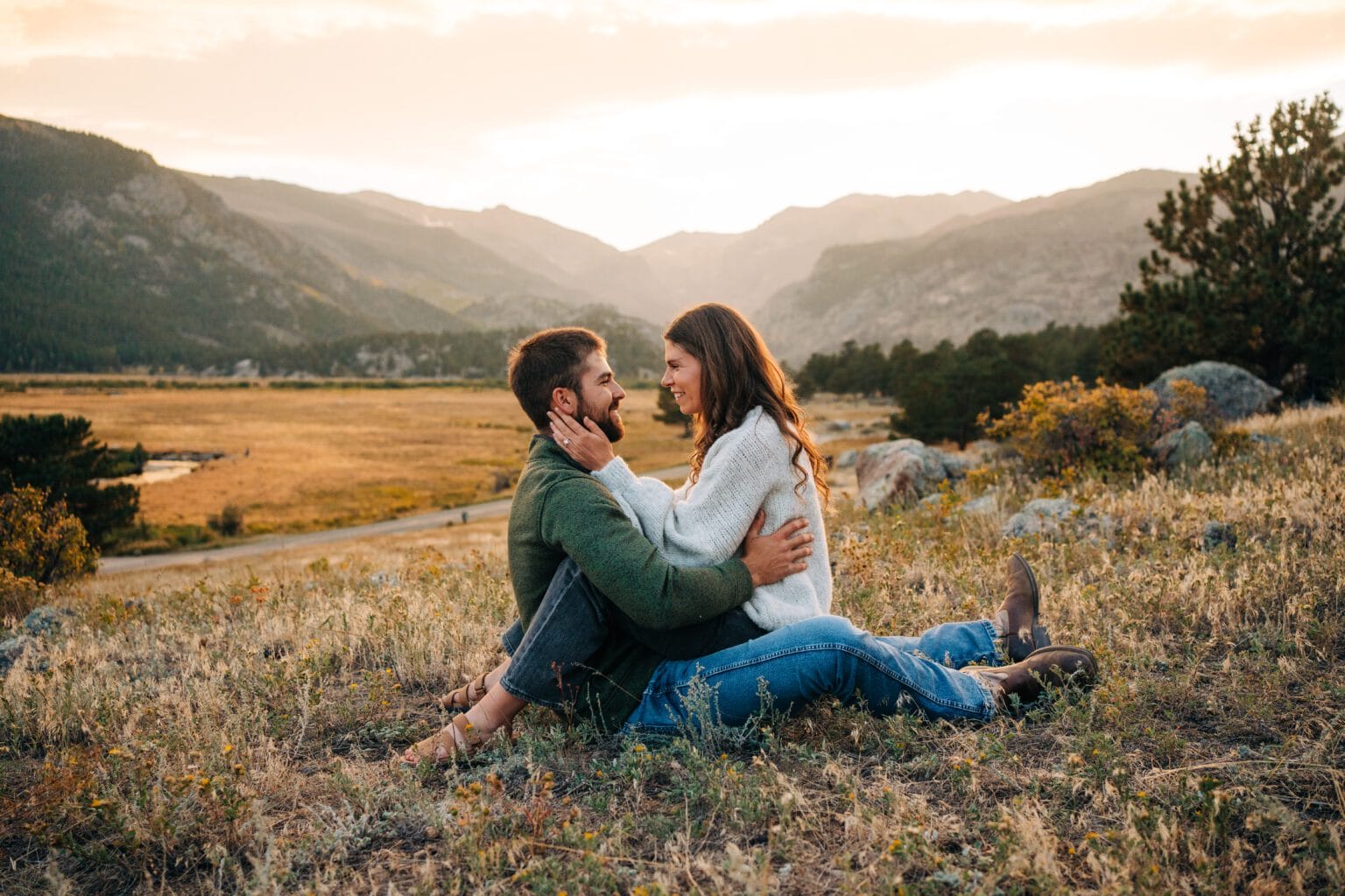 A couple sits facing each other on a grassy hillside at sunset, smiling and holding each other, with mountains and a valley in the background.