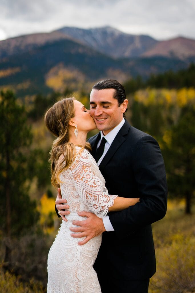 A bride in a white lace dress kisses a smiling groom on the cheek as they embrace outdoors, with pine trees and mountains in the background.