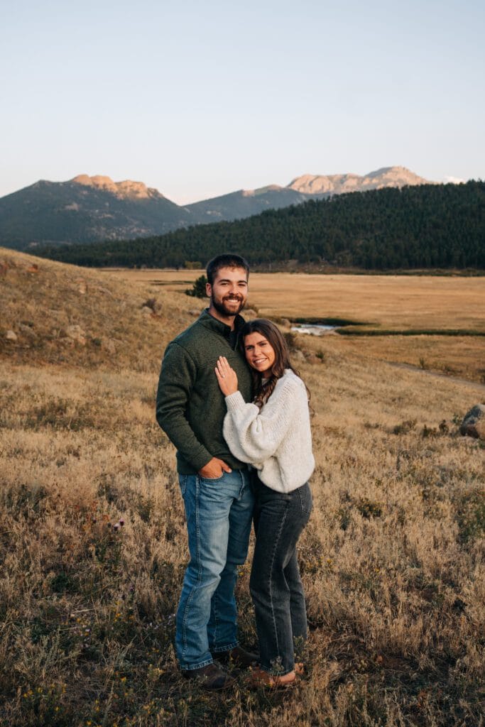 A couple stands close together in a dry grassy field, with mountains and pine trees in the background under a clear sky. The woman leans on the man, both are smiling and wearing casual sweaters and jeans.