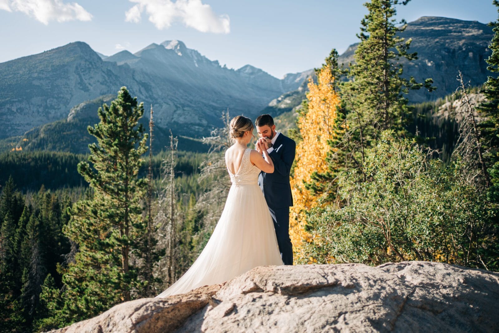 Groom raising the brides hands to kiss them as they stand on top of a rock with the Colorado mountains in the background during their Rocky Mountain National Park Elopement
