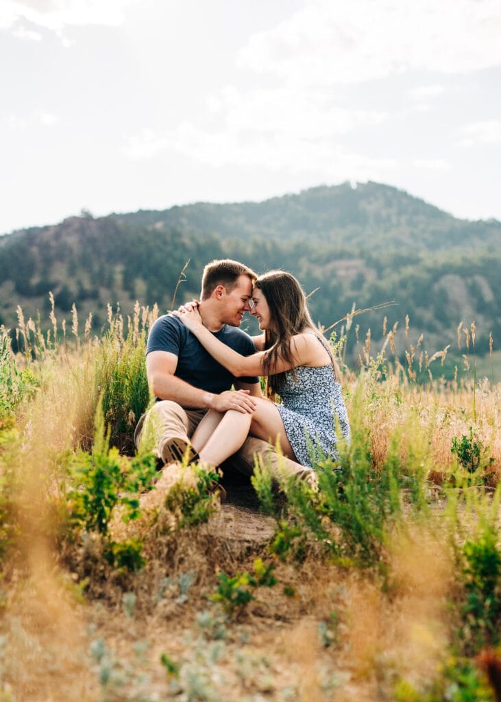 A couple sits close together in a grassy field with mountains in the background, smiling and touching foreheads affectionately on a sunny day.