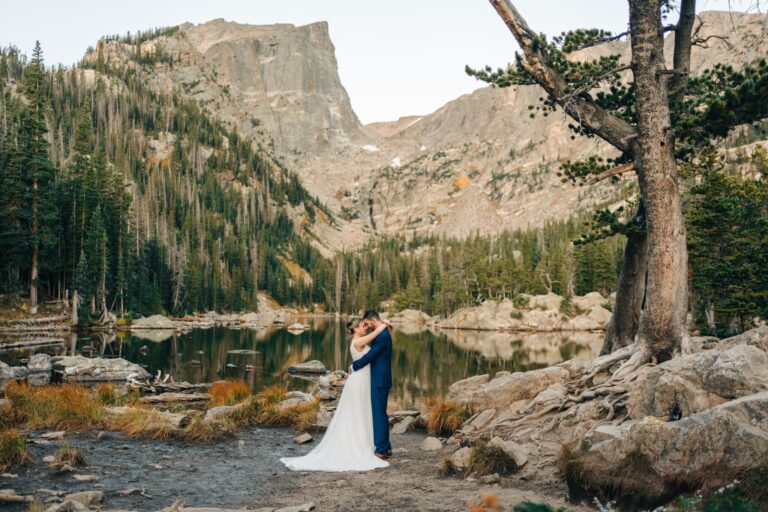 A bride and groom embrace by a calm mountain lake, surrounded by pine trees and rocky cliffs, with dramatic peaks rising in the background under a clear sky.