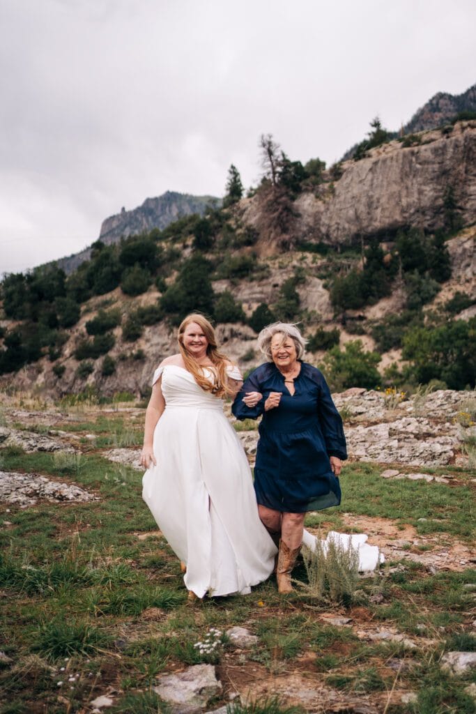 A bride in a white dress and a smiling older woman in a navy dress and boots walk arm-in-arm outdoors on rocky terrain, with green hills and cliffs in the background.