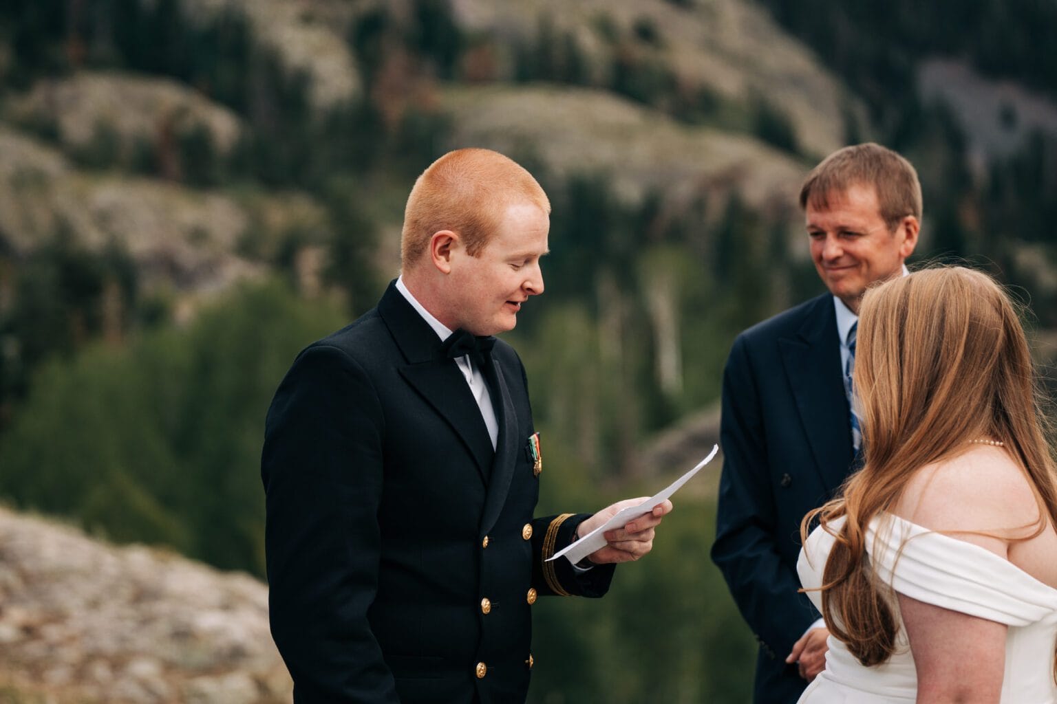 A man in formal attire reads from a paper during an outdoor wedding ceremony, with a woman in a white dress and another man in a suit standing nearby against a scenic, mountainous backdrop.