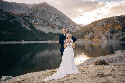 A bride and groom stand on rocky ground by a calm mountain lake at sunset, the groom holding the bride from behind as they both smile. Forested hills and rugged mountains are in the background.