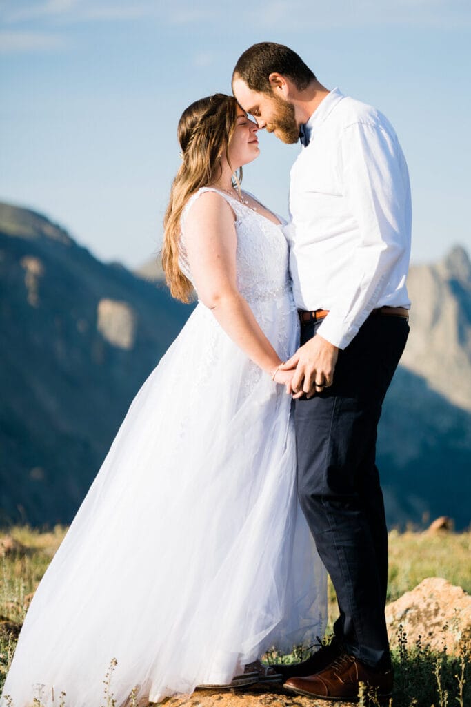 A bride and groom stand close together outdoors on a sunny day, gently touching foreheads and holding hands, with mountains visible in the background.