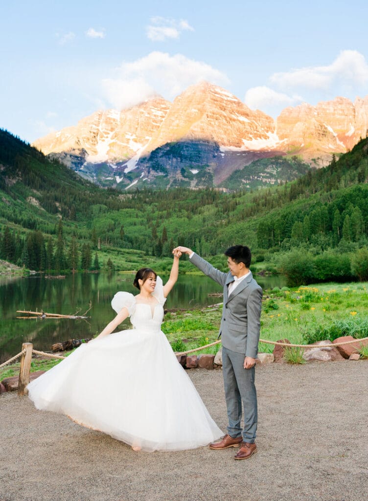 A bride in a white dress twirls with her groom in a gray suit beside a lake, with green forests and snow-capped mountains in the background under a blue sky.