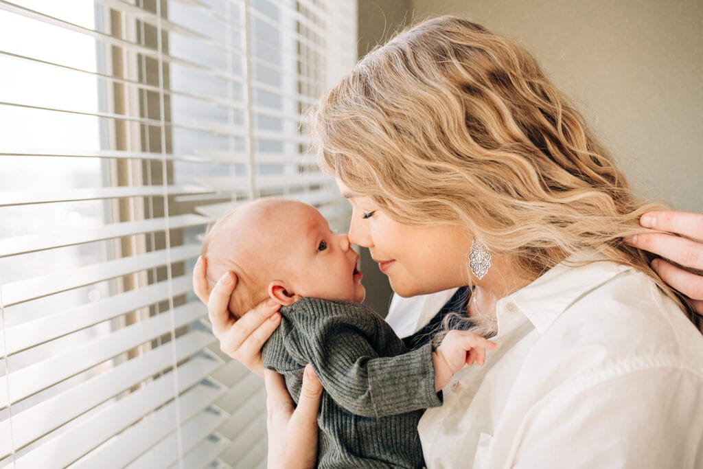 A newborn photographer captures a woman with wavy blonde hair lovingly holding her baby close, their noses touching. Sunlight streams through window blinds, creating a warm and intimate moment.