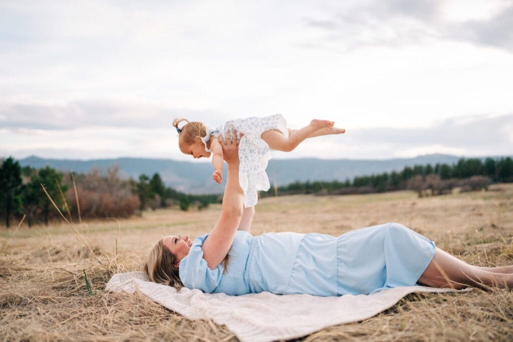A woman in a light blue dress lies on a blanket in a dry grassy field at Dawson Butte, playfully lifting a young girl in a white dress above her during their Mommy and Me Photoshoot. Trees and hills appear beneath the cloudy sky.