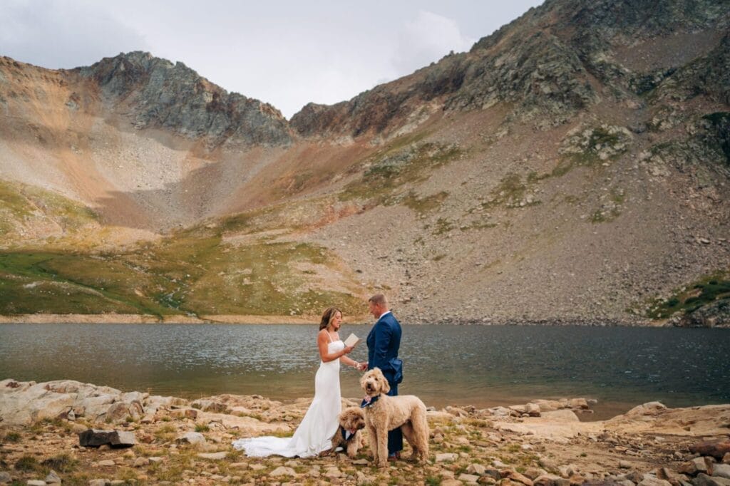 A bride and groom stand by a mountain lake, exchanging vows. Two dogs accompany them in the foreground, while rocky mountains rise under a cloudy sky in the background.