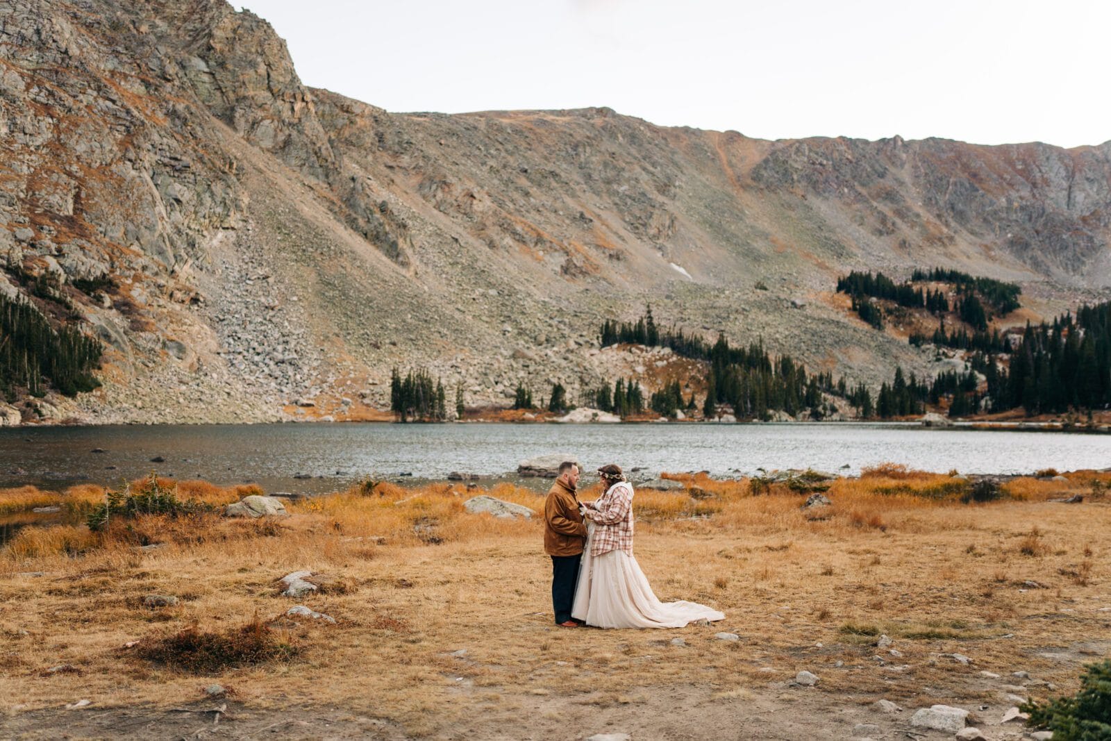 A couple stands together on dry grass by a calm mountain lake, surrounded by rocky hills and pine trees under a clear sky. The person in a dress and the other in a jacket hold hands, facing each other.