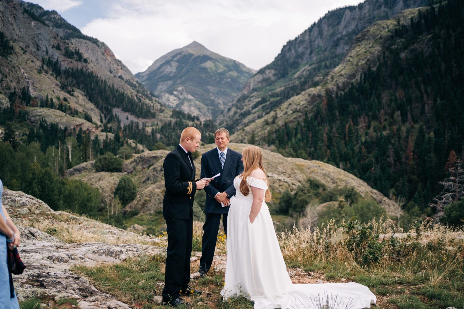 A bride and groom stand with an officiant during an outdoor wedding ceremony in a scenic mountain landscape, surrounded by green hills, trees, and dramatic peaks under a cloudy sky.