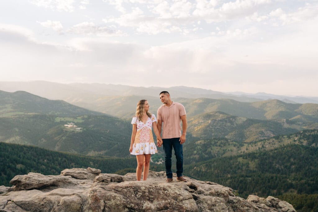 Couple holding hands on rocky cliff smiling at eachother during their sunset Lost Gulch overlook proposal in boulder colorado
