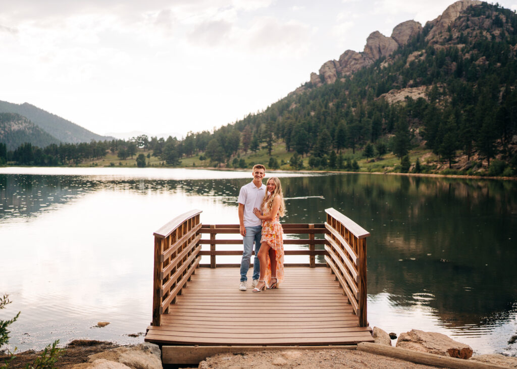 Young couple standing on a lake dock smiling at the camera during their Lily Lake Engagement Photos