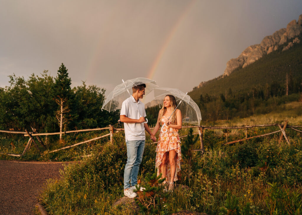 engaged couple holding hands in a meadow with umbrellas with a rainbow behind them during their lily lake engagement photos