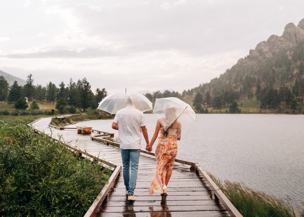 couple holding hands and walking down a boardwalk in the rain with umbrellas during their lily lake engagement photos