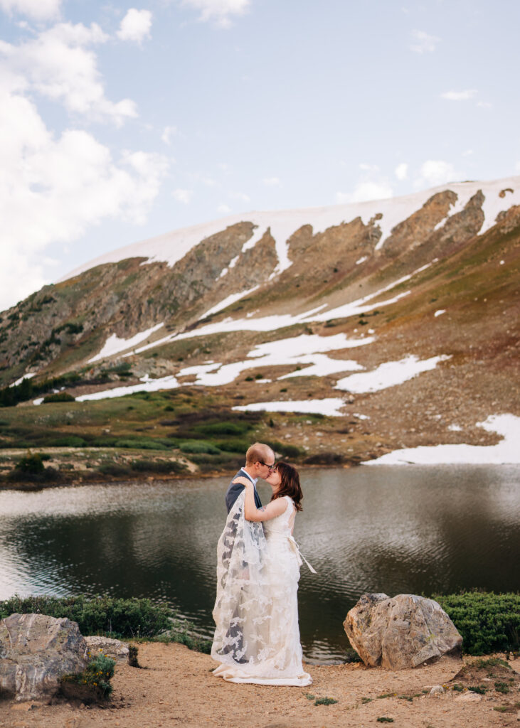 bride and groom standing by a lake with the train of her dress swept up over the grooms shoulder during their loveland pass elopement