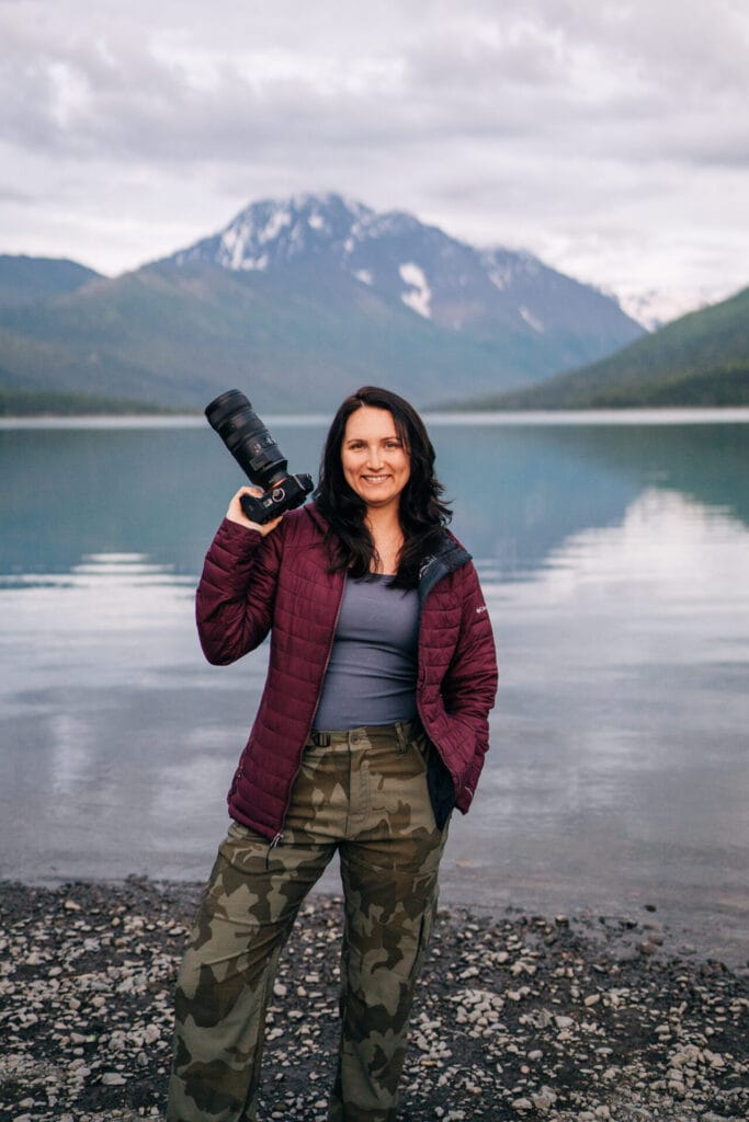 Owner of Brenna Nicole Photography standing in front of an alpine lake and mountains with her camera during her Colorado Elopement Photographer branding shoot