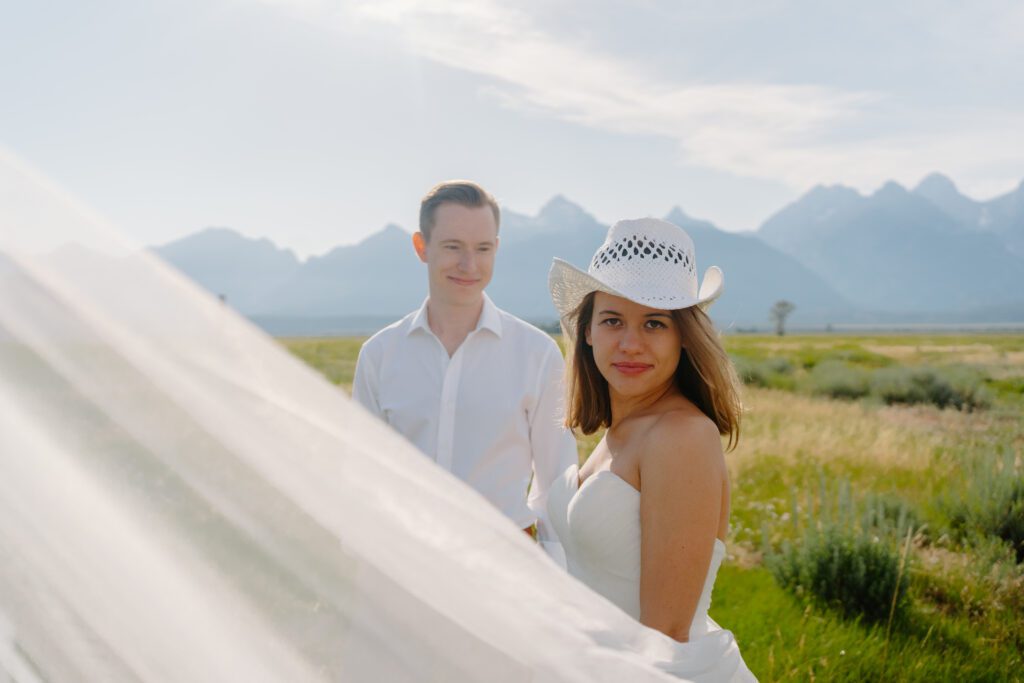 bride looking into the camera while her veil floats in the foreground and her husband looks back at her in grand teton national park during their elopement