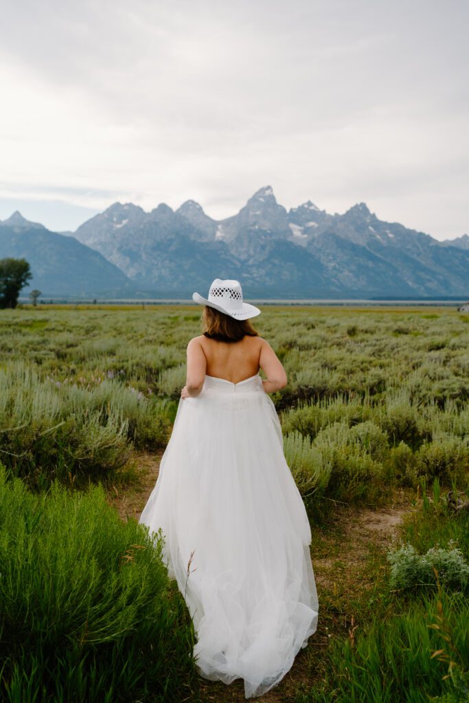 bride wearing a cowboy hat and wedding dress, walking through a field in grand teton national park during her elopement