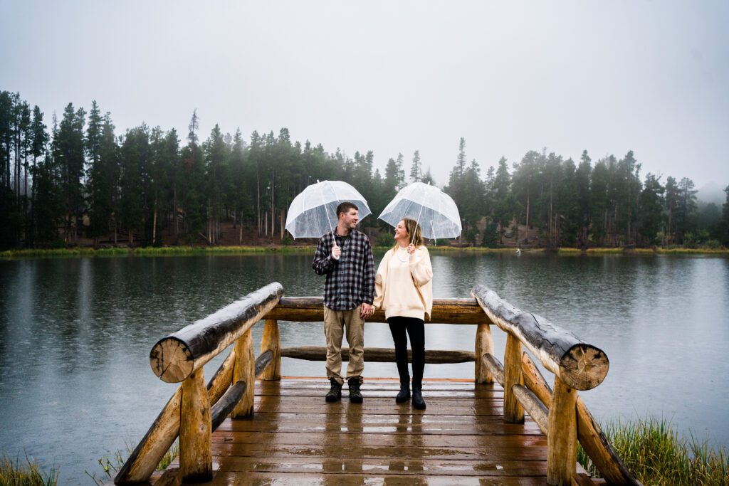 cute married couple standing at sprague lake with umbrellas in the rain during their rocky mountain national park anniversary session