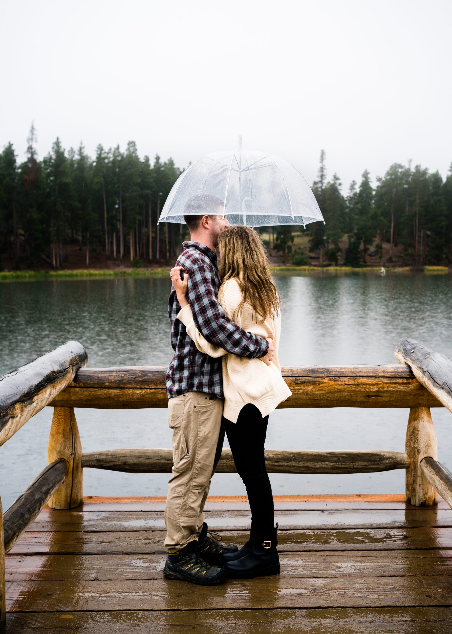 married couple standing in the rain with umbrellas at sprague lake during their rocky mountain national park engagement photos