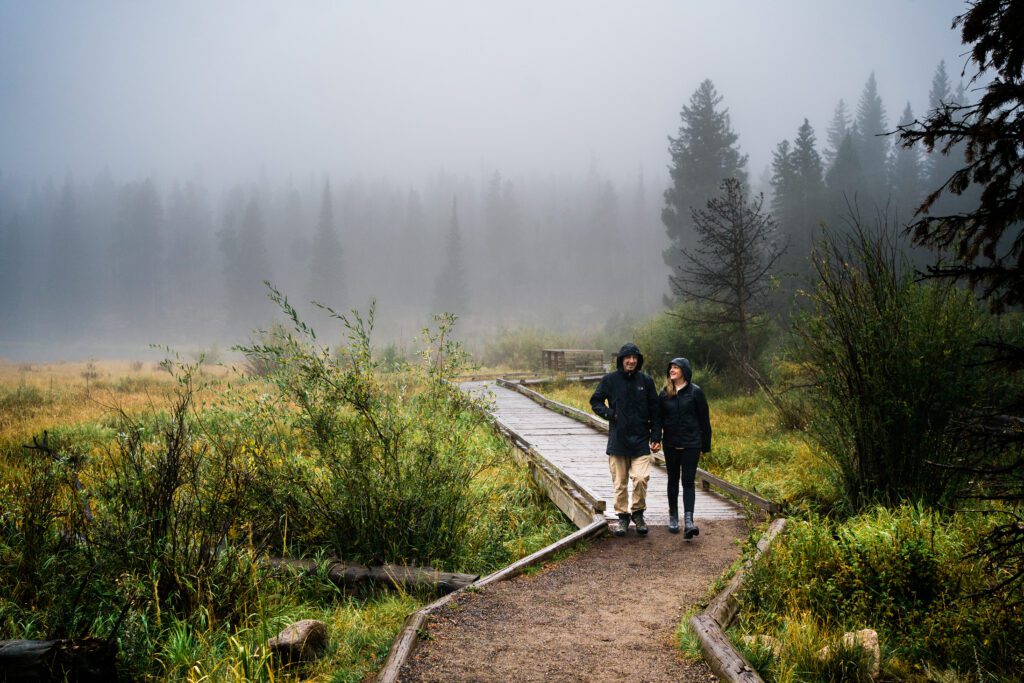 husband and wife holding hands walking through the forest in the rain during their rocky mountain national park engagement session