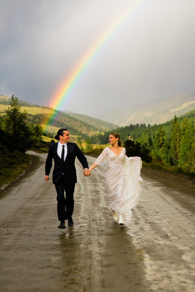 bride and groom holding hands running down the road with a huge rainbow in the background