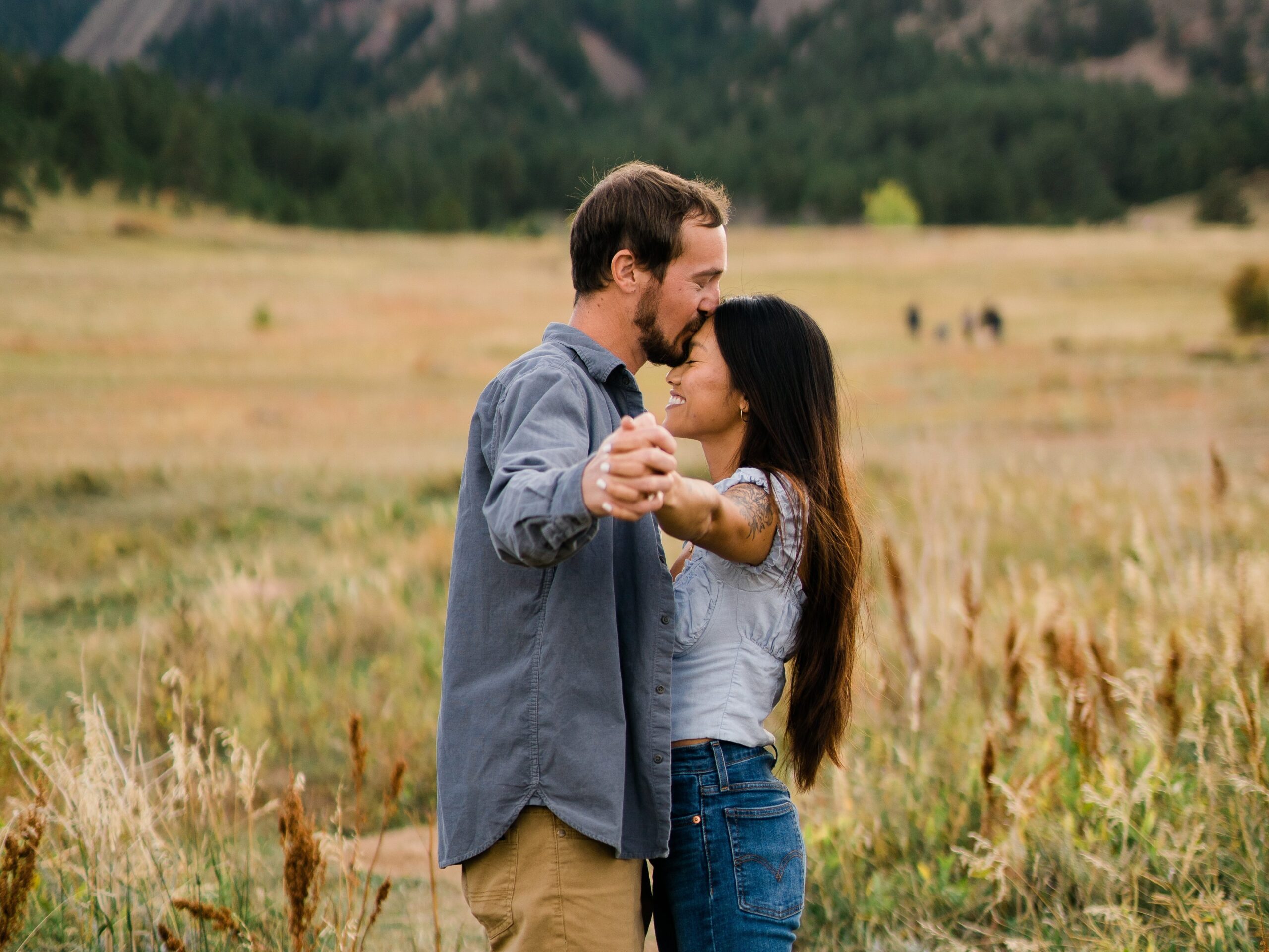 fiance kissing his girl friends forehead while they hold hands in a field during their Chautauqua Park Engagement Photos