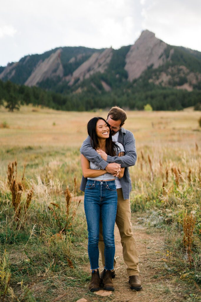 fiance kissing and hugging his girlfriend from behind in a field during their Chautauqua Park Engagement Photos
