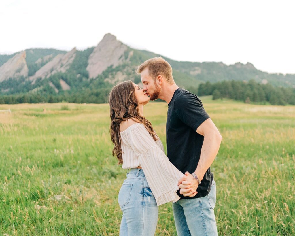 boyfriend holding his girlfriends hands as he kisses her in front of the boulder colorado flatirons during their couples session at chautauqua park