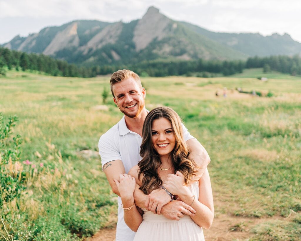 cute couple smiling at the camera in front of the boulder flatirons at chautauqua park during their colorado couples photoshoot