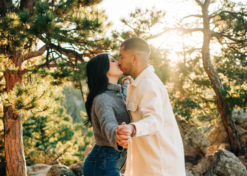 couple holding hands out towards the camera while they kiss in the background under the trees at lost gulch overlook in boulder colorado during their couples photoshoot