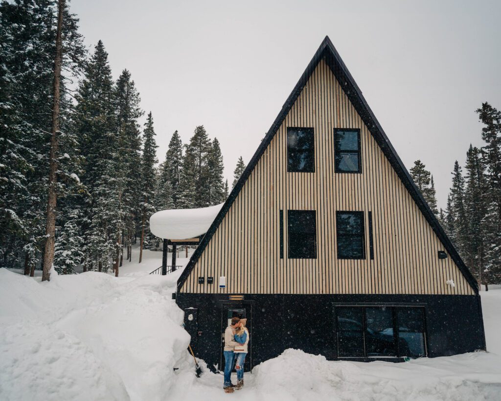 cute couple standing outside of their a-frame cabin surronded by snow and pine trees during their breckenridge engagement session