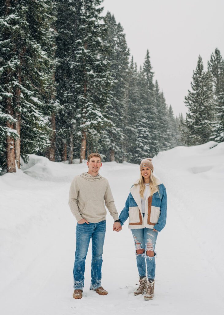 cute couple holding hands while standing on a snow covered path with pine trees in the background during their breckenridge engagement session