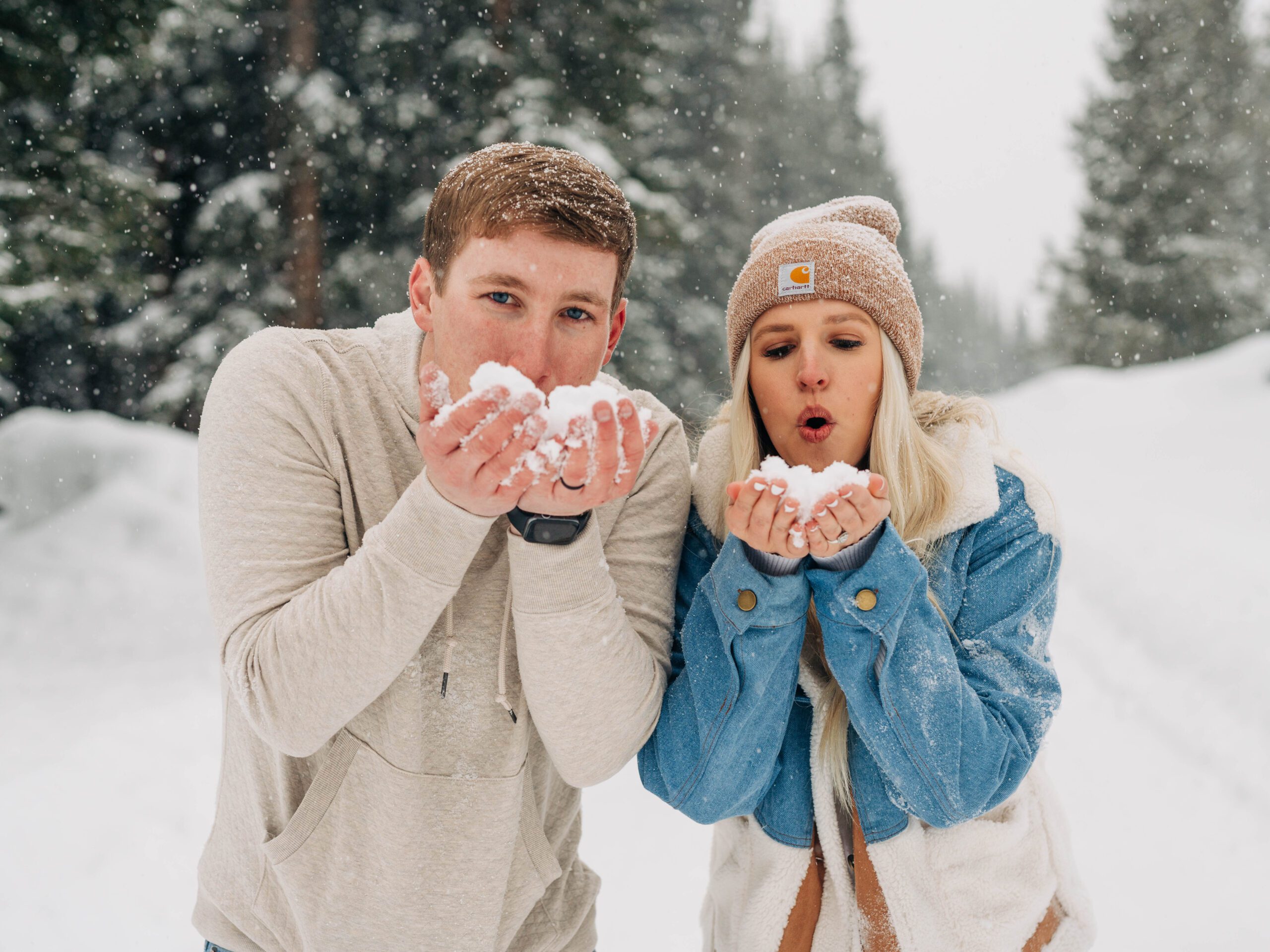 cute couple with snow in their hands blowing snow at the camera during their engagement session in breckenridge