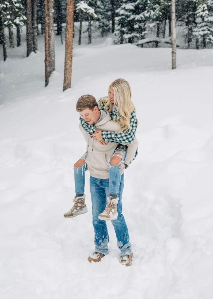 husband giving his wife a piggy back ride in the snow during their winter breckenridge engagement session