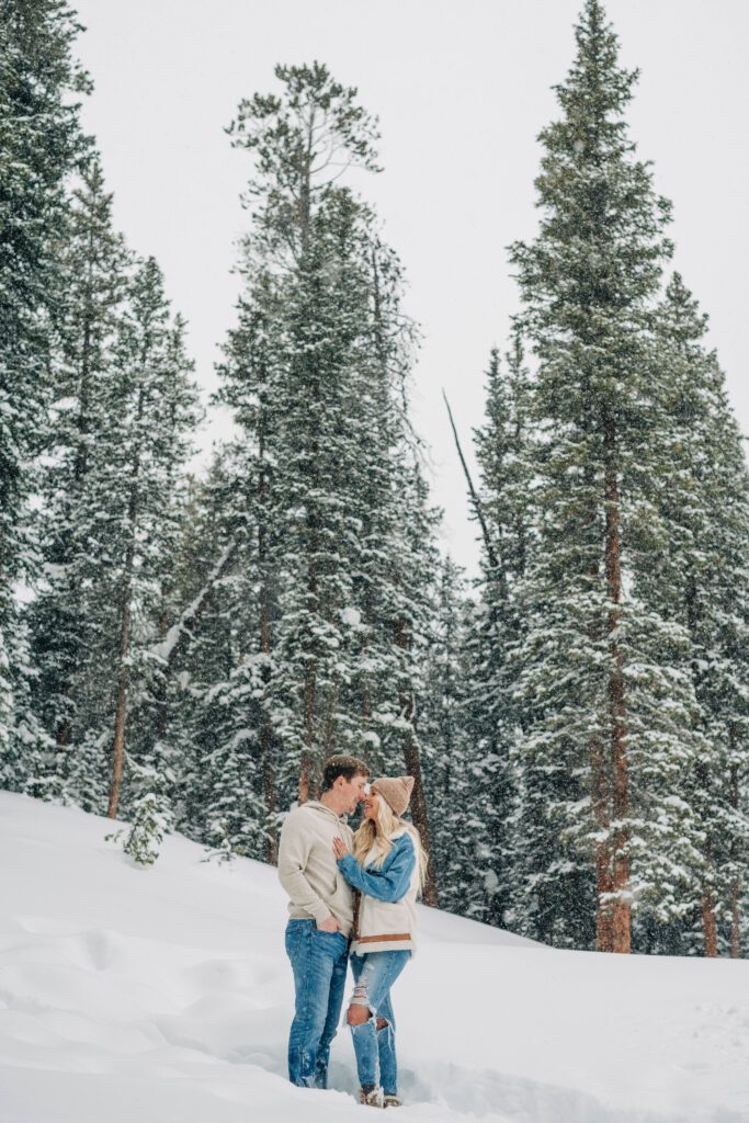 couple looking at eachother and standing in the snow with huge pine trees covered in snow in the background during their breckenridge engagement session