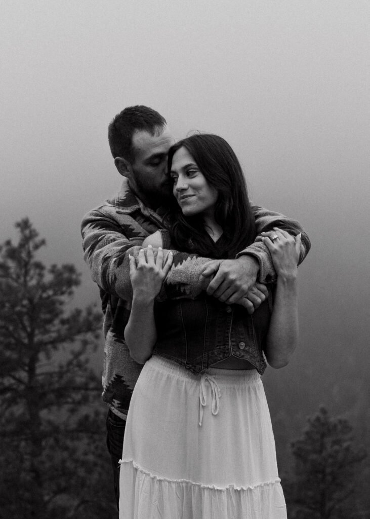 fiance hugging his girlfriend from behind on the side a misty mountain during their proposal in colorado